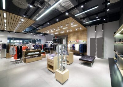 Modern clothing store interior with various clothes on racks and shelves, display cases with accessories, and bright ceiling lights. The store features a contemporary design with wooden accents.