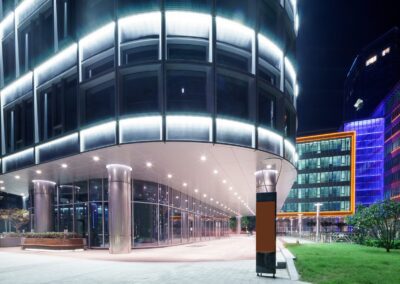 A modern glass office building illuminated at night, with a curved facade, large windows, and bright lights. An adjacent building with colorful lighting stands in the background.