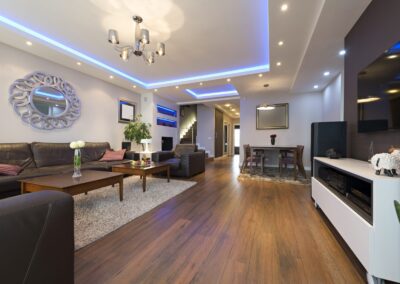 Modern living room with hardwood floors, recessed lighting, and a blue-backlit ceiling. The space includes a seating area with a sectional sofa, coffee table, wall art, and a dining area in the background.